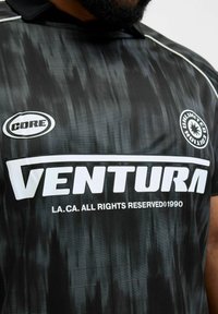 Camiseta deportiva negra con rayas verticales grises, cuello tipo polo y texto prominente en blanco que dice "VENTURA" y "CORE."
