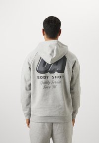 Grå hættetrøje med et stort sort grafisk print og tekst på ryggen, ribbede manchetter og kant. Blødt materiale med en justerbar hætte.