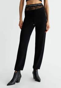 Czarne spodnie dresowe z koronkowym pasem w talii, wyposażone w sznurki i dopasowany krój. Połączone z czarnymi, spiczastymi botkami za kostkę.