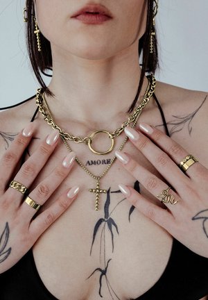 Gold jewelry features a chunky chain necklace with a circular pendant, layered with a cross necklace. Various rings adorn fingers; a tattoo is visible.
