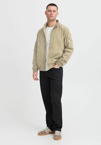 Veste bomber beige clair avec des poignets côtelés, portée sur un t-shirt blanc. Un pantalon noir à coupe droite et des baskets brunes complètent la tenue.