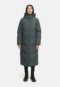 Garšs, tumši zaļš puffer mētelis ar ševronu quilted rakstu, augstu apkakli ar auklām un sānu kabatām, valkāts pār melnajām biksēm un zābakiem.