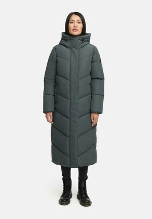 Lange, donkergroene pufferjack met een chevron-quiltpatroon, een hoge kraag met trekkoorden en zijzakken, gedragen over zwarte broek en laarzen.