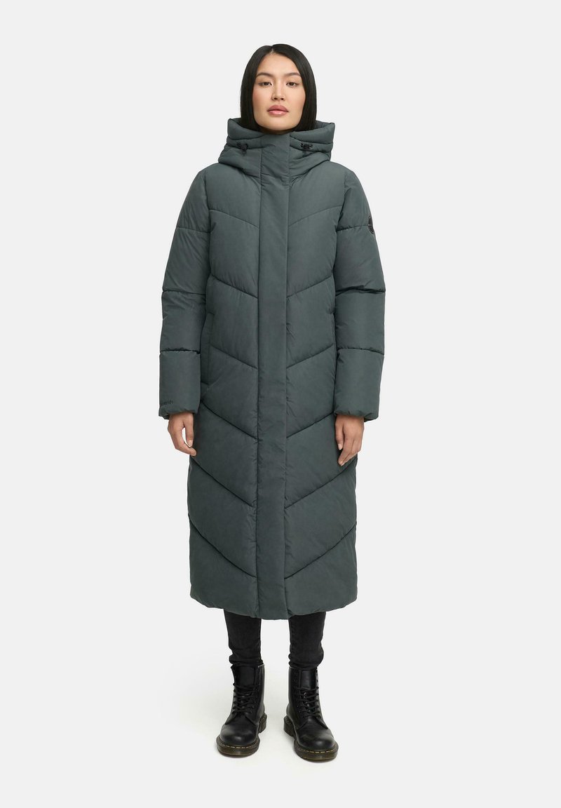 Garšs, tumši zaļš puffer mētelis ar ševronu quilted rakstu, augstu apkakli ar auklām un sānu kabatām, valkāts pār melnajām biksēm un zābakiem.