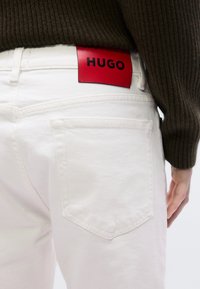 Λευκό τζιν παντελόνι με ορατές πίσω τσέπες. Μια κόκκινη δερμάτινη ετικέτα με την ανάγλυφη μαύρη επιγραφή "HUGO" είναι προσαρτημένη στη μέση. Πάνω από το παντελόνι υπάρχει υφασμένο καφέ πουλόβερ.