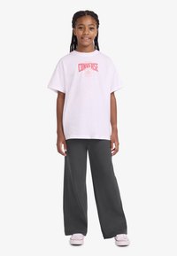 Λευκό μπλουζάκι με κόκκινο λογότυπο "Converse", που συνδυάζεται με σκούρο γκρι φαρδιά παντελόνια και λευκά παπούτσια. Το ύφασμα φαίνεται απαλό με άνετη εφαρμογή.