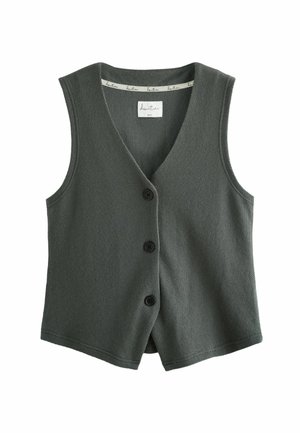Kvinde iført løs mørkegrå ærmeløs knapbluse og bukser med vide ben, stående barfodet i et minimalistisk rum med træbænk og puder.