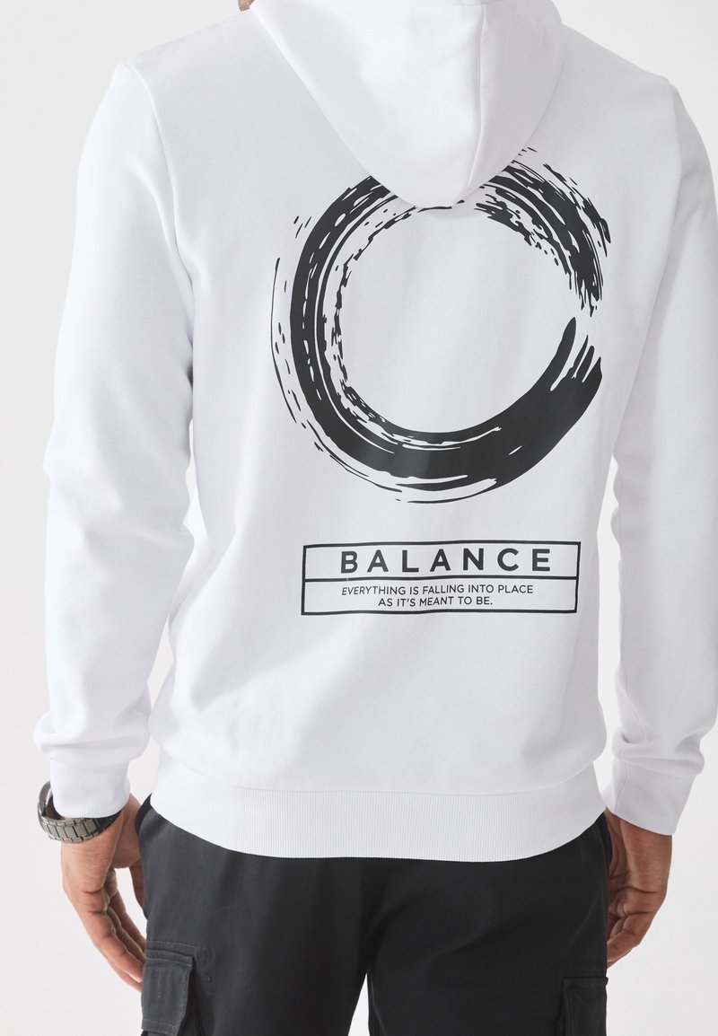 Person iført en hvid hættetrøje med et sort cirkulært penselstrøgdesign og teksten "BALANCE Alt falder på plads, som det skal være."