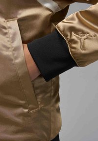 Giacca bomber in satin dorato con polsini a coste neri. Presenta una tasca laterale e un dettaglio del logo ricamato sulla manica. Texture liscia.