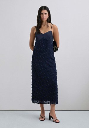Vestido sin mangas de color azul marino con un corsé ajustado y un patrón de puntos texturizados y en relieve en la falda. Largo hasta el suelo y de forma recta.