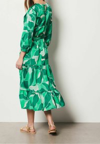 Kvinna som bär en grön blommig mönstrad midiklänning med långa ärmar och beige platta sandaler, stående mot en ljus vägg.