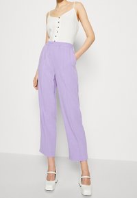 Haut blanc sans manches avec des boutons, assorti à un pantalon lavande à jambes larges. Le pantalon dispose d'une taille élastique et de poches latérales. Le modèle porte des chaussures à plateforme blanches.