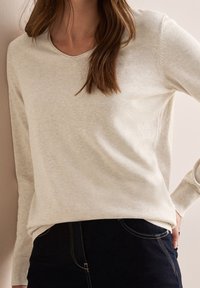 Pull en maille beige clair à manches longues avec un col rond. Texture douce et coutures discrètes. Associé à un jean noir avec des surpiqûres jaunes.