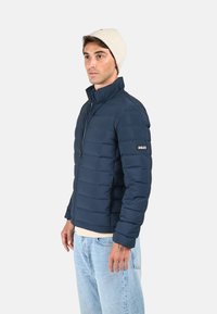Doudoune bleu marine avec des coutures horizontales, un col montant et une fermeture éclair. Portée par-dessus une chemise claire et assortie à un jean clair.