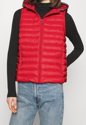 Mujer con chaleco acolchado rojo sin mangas con capucha, camiseta negra de manga larga y vaqueros azul claro, de pie con una mano en la cadera.