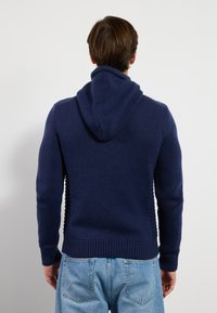 Marinblå stickad hoodie med en texturerad yta, ribbade manschetter och nederkant, samt en stor, lös huva. Bakifrån visas inga ytterligare detaljer.