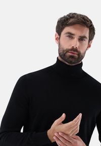 Schwarzer Rollkragenpullover, taillierte Passform, gestrickte Textur, gerippter Kragen, lange Ärmel, getragen mit verschränkten Händen, neutraler Hintergrund.