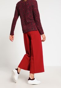 Røde brede bukser parret med en tekstureret sweater i mørkerød og sort mønster, komplementeret af hvide sneakers med burgunderrøde accenter.