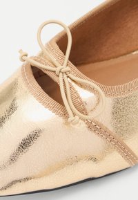 Zapato plano de material sintético dorado brillante con una superficie texturizada. Presenta un ribete de tela beige y un lazo decorativo en la parte delantera.