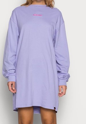 Osoba w luźnej, lawendowej sukience z długimi rękawami, z różowym logo "ellesse" na piersi, stojąca na jednolitym tle.