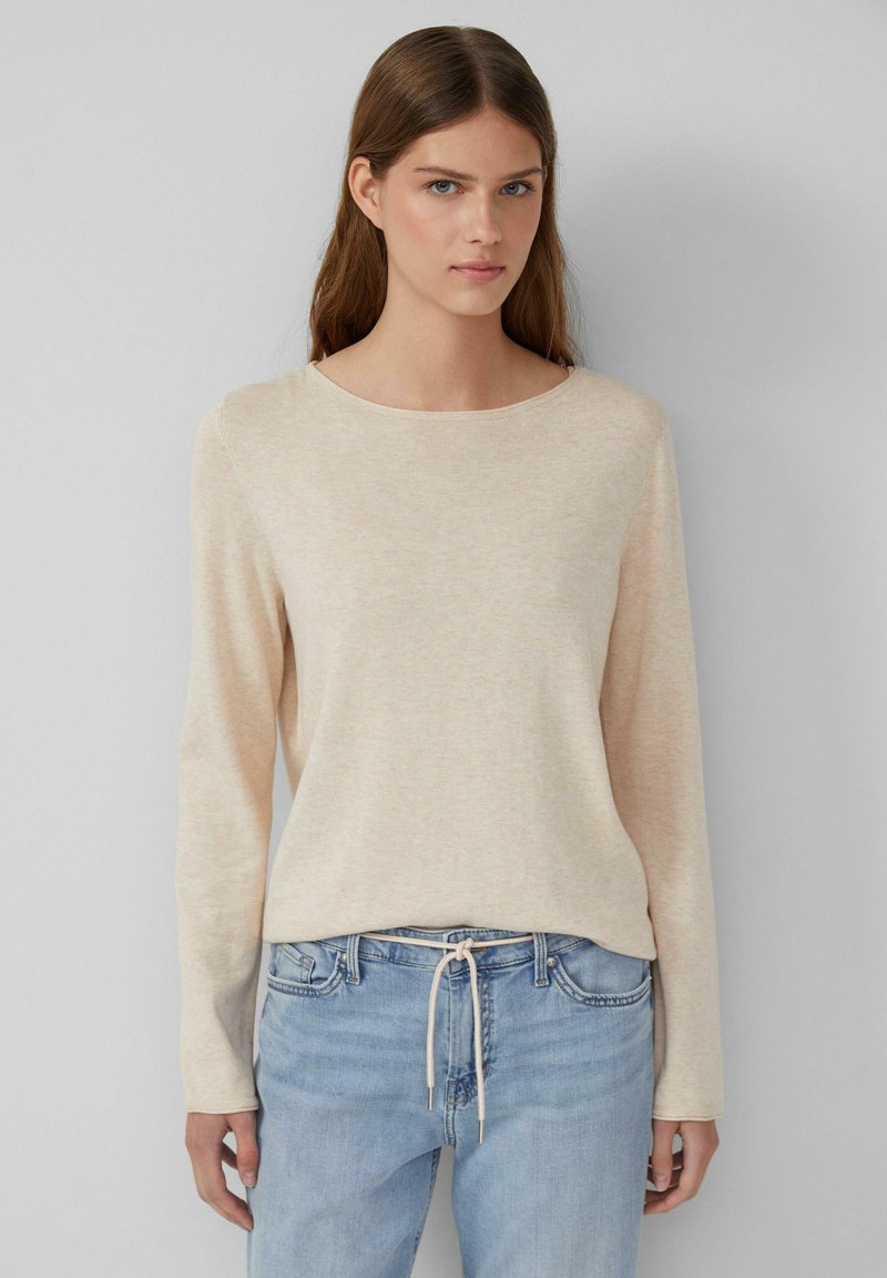 Maglione a maniche lunghe color beige chiaro con una consistenza morbida e un collo rotondo, abbinato a jeans in denim azzurro chiaro con vita regolabile.