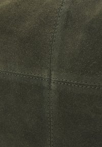 Gros plan sur un matériau en daim vert foncé avec des coutures apparentes formant un motif en croix.
