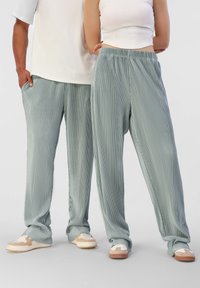 Pantalons plissés vert clair en matériau doux, avec une taille élastique et des poches latérales, associés à des baskets beiges et crème.