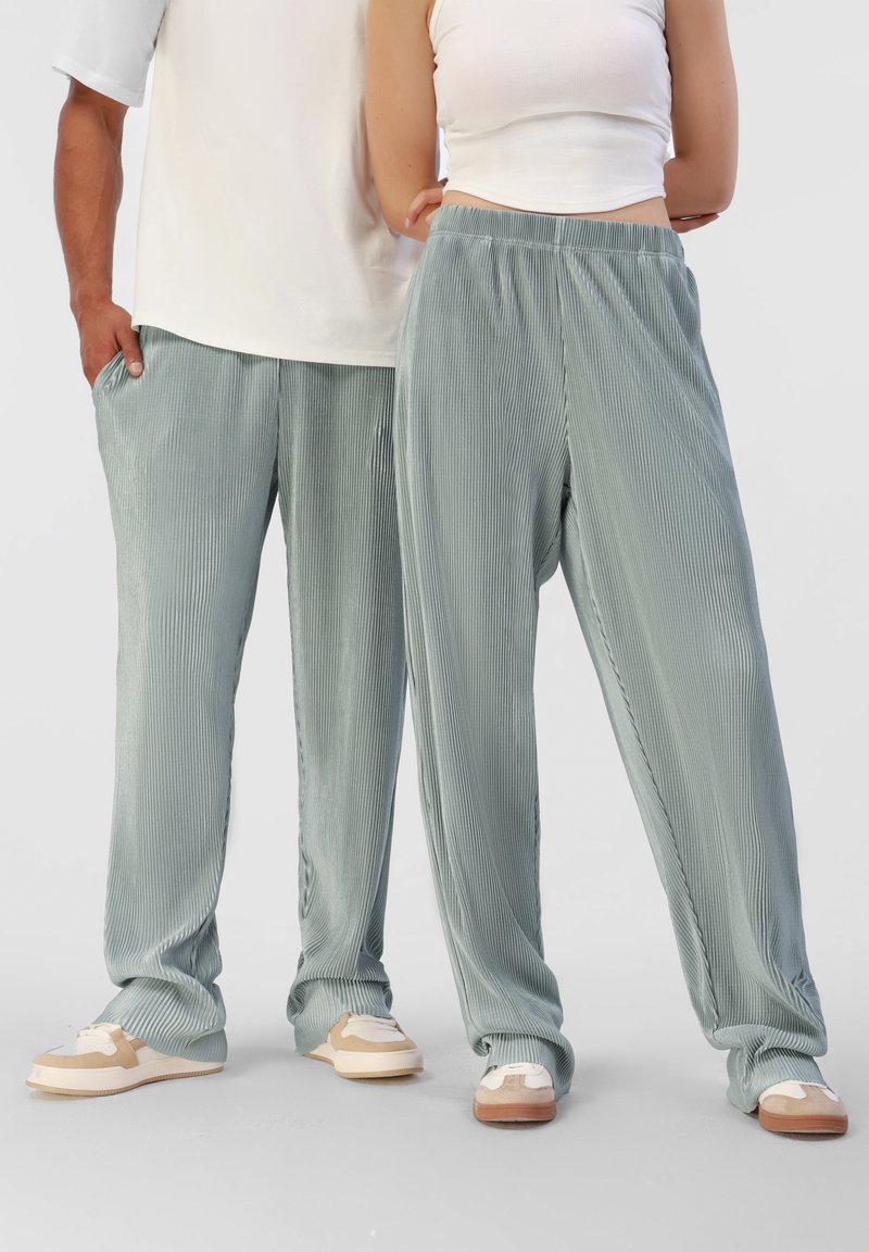 Pantalons plissés vert clair en matériau doux, avec une taille élastique et des poches latérales, associés à des baskets beiges et crème.