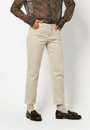 Person wearing beige straight-leg pants, brown patterned long-sleeve top, beige socks, and dark brown tassel loafers, standing with hands in pockets.