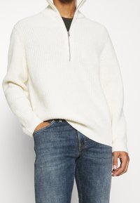 Pull tricoté couleur crème avec col demi-fermeture éclair, nervures texturées, coutures d'épaule tombantes, porté avec un jean bleu.