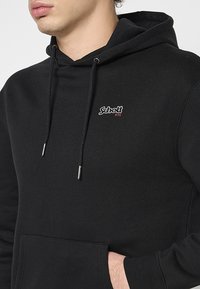 Felpa nera in cotone con tasca anteriore, lacci e un piccolo logo bianco e rosso sul petto. Tessuto morbido, vestibilità comoda.