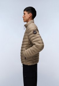 Beige pufferjakke med horisontalt quiltet mønster, høj krave, lynlåslommer og cirkulært logo-patch på venstre ærme.