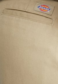 Beżowa, teksturowana tkanina z kieszenią z tyłu i naszywaną metką z logo Dickies. Materiał wygląda na mocny i trwały.