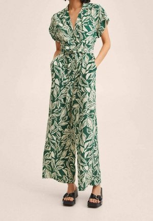 Kvinde iført grøn jumpsuit med vide ben med hvidt bladmønster og sorte platformssandaler, hænderne i lommerne.