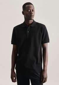Zwarte polo in katoen met een klassieke kraag, korte mouwen en een discreet logo op de borst. Glad textuur, slim fit.