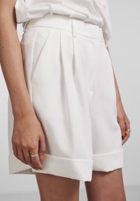 Bílé plisované šortky vyrobené z hladké látky, s ohrnutým lemem a přiléhavým pasem, bočními kapsami a jemným leskem.