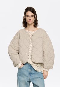 Quilted beige jacket with snap buttons, rounded shape, and oversized sleeves, layered over a lace top and denim jeans.