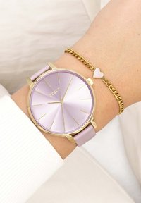 Montre violette avec un cadran rond, accents dorés et un bracelet rose, associée à un bracelet en chaîne doré avec un petit charme en forme de cœur.