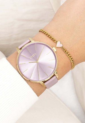 Montre violette avec un cadran rond, accents dorés et un bracelet rose, associée à un bracelet en chaîne doré avec un petit charme en forme de cœur.