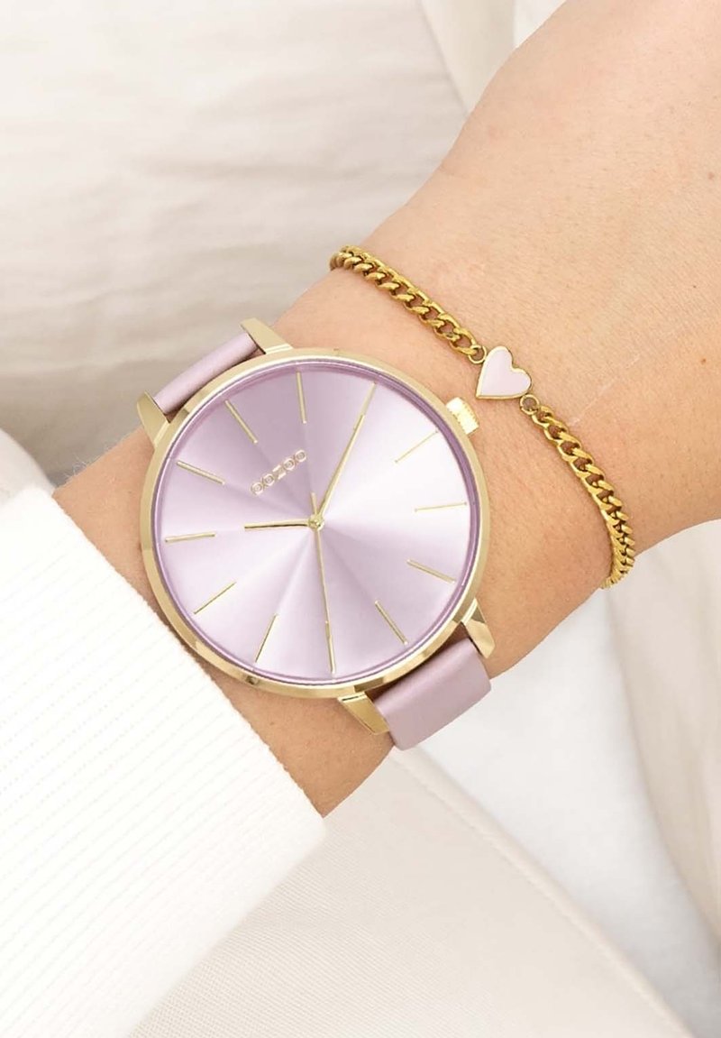 Montre violette avec un cadran rond, accents dorés et un bracelet rose, associée à un bracelet en chaîne doré avec un petit charme en forme de cœur.