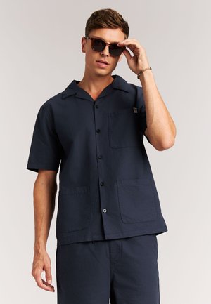 Marineblauw overhemd met korte mouwen gemaakt van gestructureerde stof, voorzien van een kraag, knoopsluiting en twee zakken aan de voorkant. Draagt een zonnebril.