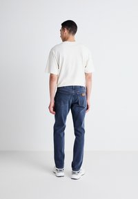 Pantalones vaqueros de mezclilla azul oscuro con un ajuste slim, que presentan costuras visibles y un parche de cuero marrón en la parte posterior. Combinados con una camisa beige clara.
