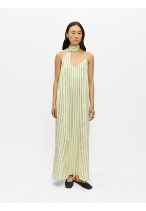 Femme debout portant une robe longue maxi sans manches vert pâle à fines rayures verticales et une longue écharpe assortie, associée à des chaussures plates noires.