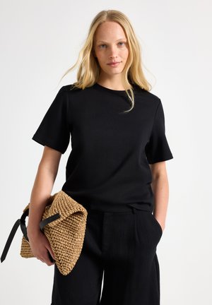 Lyshåret kvinde iført sort kortærmet skjorte og bukser, der holder en vævet beige clutchtaske med sort rem, stående mod en ensfarvet baggrund.