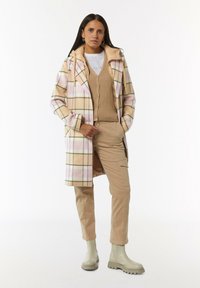 Geruite jas in zacht beige en roze, met een rits aan de voorkant en een oversized kraag. Gecombineerd met een ribbeltop in beige en kaki broek, en groene laarzen.