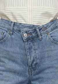 Jean en denim bleu taille haute avec une taille asymétrique boutonnée, porté avec une chemise beige clair à fines rayures rentrée.