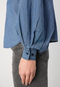 Lys blå denimskjorte med løs passform, lange puffermer og to-knappe mansjetter. Stoffet har en strukturert vev. Brukt med grå bukser.