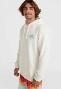 Cream-colored hooded sweatshirt with a front pocket, featuring a turquoise circle logo. The wearer has patterned shorts in red, blue, and yellow.