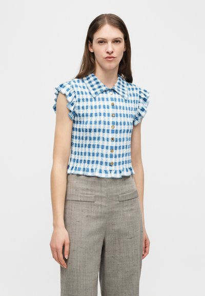 Γυναίκα που φοράει μπλε και λευκό κοντομάνικο καρό πουκάμισο με κουμπιά και βολάν στους ώμους, συνδυασμένο με γκρι ψηλόμεσο παντελόνι.