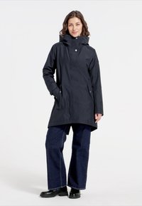 Mörkblå vattentät jacka med hög krage, tryckknappar och sidofickor. Kombinerad med vida mörka jeans och svarta stövlar.